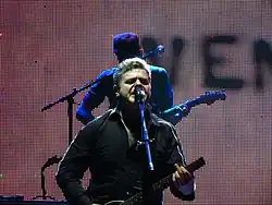 A man with grey hair sings into a microphone, playing a string instrument