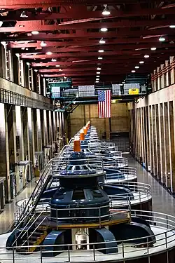 A picture looking down on eight generators in a line receding away from the viewer. Each generator is a cylinder with dark red walls, with a railing and walkable space on the white top of each generator. On top of each generator is a blue structure that comes to a point. On the very top of each generator is are white lights with black letters "N1" (closest) through "N8" (furthest). All are lit except for "N4". There are access catwalks on top of each generator. The generators are in a large stone hallway with metal beams in the ceiling. There are two gantry cranes at the far end of the room.
