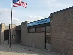 A one-story brick building in a 1960s architectural style stands with a waving American flag before it.