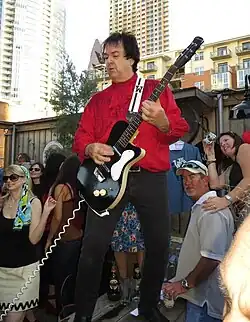 Jeff Walls, a plus sized caucasian man wearing a red shirt, plays guitar in an outdoor venue