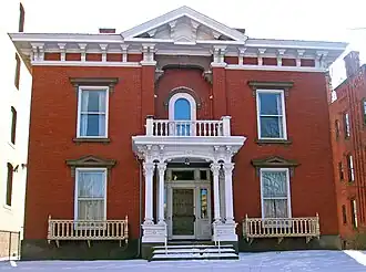 A brick house with white ornate wooden trim seen from the front. Its roof has a small pointed section in the middle, and the front door is boarded over. There is snow on the ground in front and the steps.