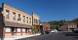 A streetscape of two-story brick buildings with angled parking in front and a forested ridge in the background