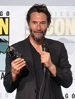 Keanu Reeves looks at the camera while holding a microphone and a trophy