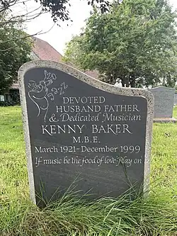 A gravestone with slate inset with slightly overgrown grass at the base and other gravestones in the background. The gravestone reads "Devoted Husband, Father and Dedicated Musician. Kenny Baker M.B.E. March 1921 to December 1999. If music be the food of love. Play on." There is also a line drawing of a bald man playing a trumpet with the words 'Jazz' next to it.
