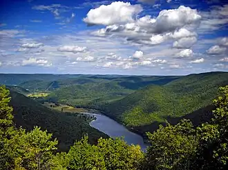A view from atop a mountain of a blue lake between roling green hills and low mountains under a partly cloudy sky