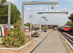 A man with a white shirt and white pants sitting on a brown bench facing the left with another bench behind him all under a light blue sky with white clouds