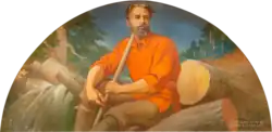 Man in pull over cotton shirt holding staff sitting on cut logs. Behind him is a river with logs piled up and sticking out as if in a log jam.