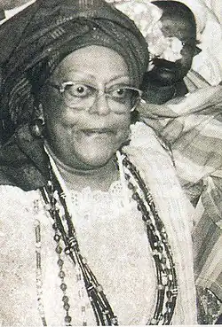 a black and white headshot of Mãe Cleusa Millet wearing a headdress, glasses, beaded necklaces, and white clothing