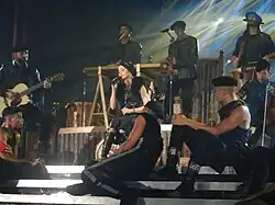 Madonna sitting onstage singing, surrounded by her dancers in a circle.