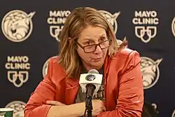 Portrait of a woman in her mid fifties holding a microphone with shoulder length highlighted hair, wearing silver hoop earrings, a Sylvia Fowles tribute t-shirt, gray-patterned suit jacket