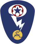 Oval shaped shoulder patch with a deep blue background. At the top is a red circle and blue star, the patch of the Army Service Forces. It is surrounded by a white oval, representing a mushroom cloud. Below it is a white lightning bolt cracking a yellow circle, representing an atom.