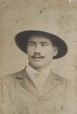 black-and-white image of Mario Tamagno wearing a light-coloured outfit, seated in a chair, looking just right of camera