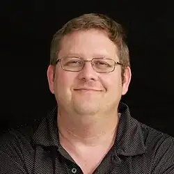 a white man with short brown hair wearing a black polo and rectangular glasses smirks
