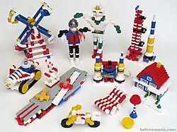A photograph of multiple toys constructed from interconnecting plastic blocks of various colors. They depict a Ferris wheel, the Japanese television characters Kamen Rider and Silver Kamen, a rocket ship and launch station, a moon rover, a battleship with two planes, an electronic clock, a house, a beach chair, a lamp and table, and a bicycle.