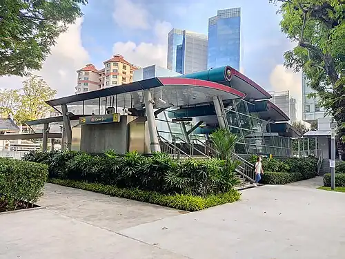Exit A of the station, with a curvilinear roof structure. A staircase leads up to the entrance, flanked by lush greenery and well-maintained shrubs.