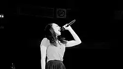 A black-and-white image. Against a black background, a young woman in side profile holds a microphone high and belts into it.