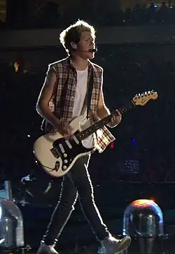 A young adult male, singer Niall Horan, on stage playing an electric guitar as part of One Direction during their Where We Are Tour. He wears a sleeveless plaid shirt over a white tank top. The plaid shirt is unbuttoned, revealing the tank top underneath. He also wears jeans.