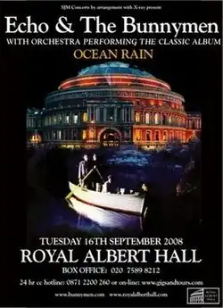 An poster advertising a concert with a black background and a picture of a circular building. Superimposed on the picture is a rowing boat with four men; two men are stood side by side at the back of the boat each holding an oar, the third man is sat in the centre of the boat and the fourth man is leaning over the front of the boat with his hand in the water. White text is on the poster at the top and the bottom giving details of the concert.