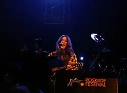 A woman sat down playing a guitar and singing into a microphone