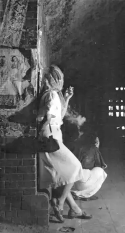 A woman stands in a tunnel smoking, another woman squats beside her.