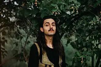 Qais Essar wearing an embroidered vest over a black long-sleeved top, looking directly at camera, with foliage in background