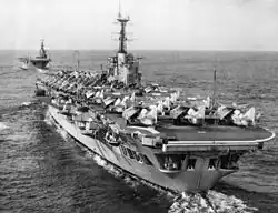 Photograph of an aircraft carrier from behind. Numerous aircraft with their wings folded are sitting on the flight deck. A second aircraft carrier is in the background, leading the first.