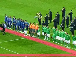 Several men standing on a grass football field. There are twenty five men standing in a straight line; eleven wearing blue on the left, three wearing orange tops and black shorts in the middle, and eleven wearing green on the right. In front of this line is a long narrow piece of red carpet. Behind the line are several middle aged or elderly men, wearing black and playing a variety of brass and percussion musical instruments.