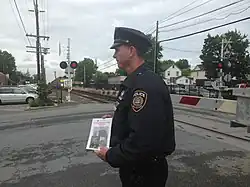 A police officer wearing a dark blue uniform faces left, holding a white pamphlet. Behind him a grade crossing gate is down, red lights are on and a train is approaching