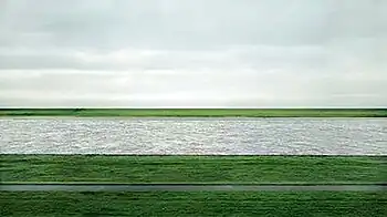 A photograph of the Lower Rhine river flowing horizontally through green fields under an overcast sky in Germany