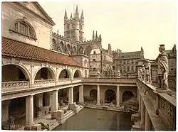 A late-nineteenth-century Photochrom of the Great Bath at the Roman Baths. Pillars tower over the water, and the spires of Bath Abbey – restored in the early sixteenth century – are visible in the background.