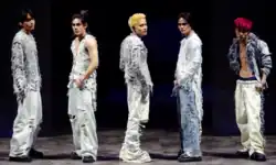 A group photo of SB19 onstage facing towards the camera, each wearing light-colored outfits