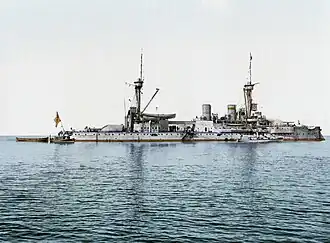 A large gray battleship with two tall masts sits idly in calm waters. Three small boats are tied alongside.