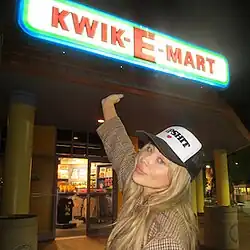 A woman with long, blonde hair showing her hand to a store with a neon sign reading 'Kwik-E-Mart', wearing a black and white cap reading 'Dipshit'.