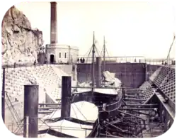 A photograph with two steamships resting in a dewatered drydock with a building housing the engine for operating the lock's gates in the background