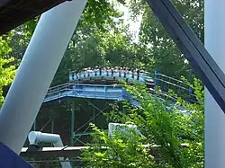 A SooperDooperLooper train is depicted as cresting one of its various hills with passengers aboard. Trees and foliage cover the background and foreground of the roller coaster.
