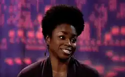 A woman with tall black hair and a wide smile wears a black jacket and looks to the right.