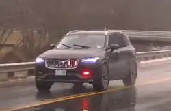 A new, black Volvo SUV driving in the rain, with red emergency lights activated while a firefighter responds to a call