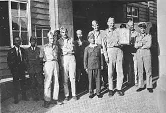 A group of men in uniforms stand in front of a wooden building. One, holding a box, is very tall, towering over the Asian man next to him.