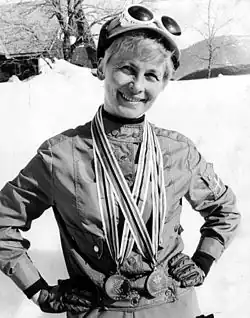 A smiling short-haired blond woman wears a track suit with gloves, and a hat with white skiing goggles. She holds her hands on her waist and has two medals around her neck. Behind her, a sunny and snowy landscape.