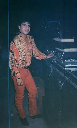Tony Barrera stands in front of the DJ booth at a Polymarchs show posing for a photo wearing a flamboyant and bright orange outfit.