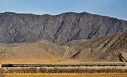 Railway line near Spezand in Dasht Tehsil, showing a train moving across a mountainous landscape dominated by rocky slopes and sparse vegetation.