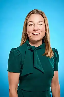 Mayor Lauren McLean smiling in a professional headshot. She is wearing a dark green short-sleeved dress with a bow tie at the neck. Her straight, shoulder-length blonde hair is worn down. The background is a solid bright blue, giving the image a clean and polished appearance.