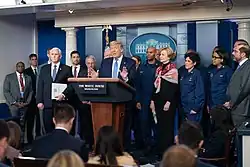 Trump speaks in the West Wing briefing room with various officials standing behind him, all in formal attire and without face masks