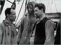 Three men in well-worn clothing stand on the deck of a ship. Man on right is thickset, bearded, with cigarette in mouth. Man in centre is hooded; man on left, also bearded and with receding hair, is shown right profile.