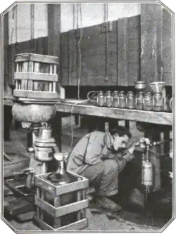 A man in workwear kneels under a table with many jars, surrounded by pipes and jugs