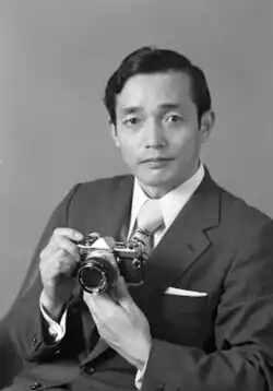 Black and white portrait of Yoshihisa Maitani, a camera designer who spent his entire career with Olympus. Mr. Maitani is pictured holding a small camera while wearing a suit and tie.