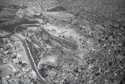 Aerial photograph of a city: the large rock of the Acropolis with the ruins of the Parthenon temple at centre; the Agora is an emptier area to its northwest.