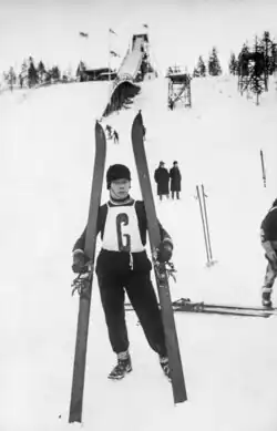 A man holding skis stands on a snow-covered mountain with other people in the background in a black and white photo.