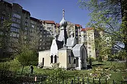 Современный облик часовни, окруженной цветниками и газоном, на фоне многоэтажного дома