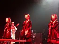 YUIMETAL, SU-METAL, и MOAMETAL в чёрных кимоно с лицами, закрытыми маской лисы, выступая на сцене